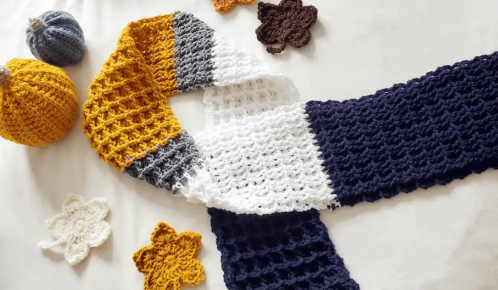 A crochet scarf with large chunks of color, the bottom of the scarf being dark blue, with a layer of white above it, a small layer of gey, and then a band of yellow in the middle of the two sides.