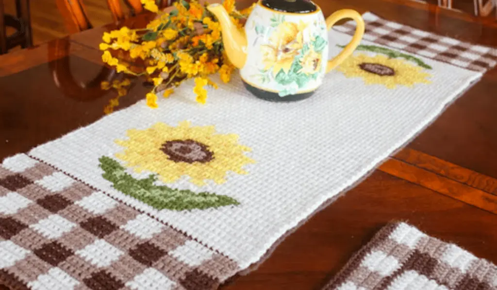 A sunflower crochet table runner with gingham rows at the bottom in white an brown.