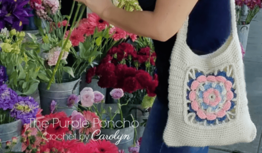 A crochet tote bag with a long strap and a prominent flower design on the side. The body of the bag is all white yarn.