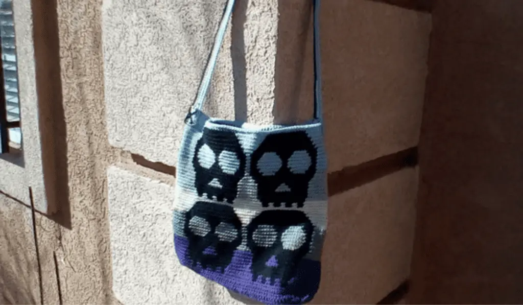 A crochet bag pattern with four skulls on the side and rows of purple, grey, and white yarn.