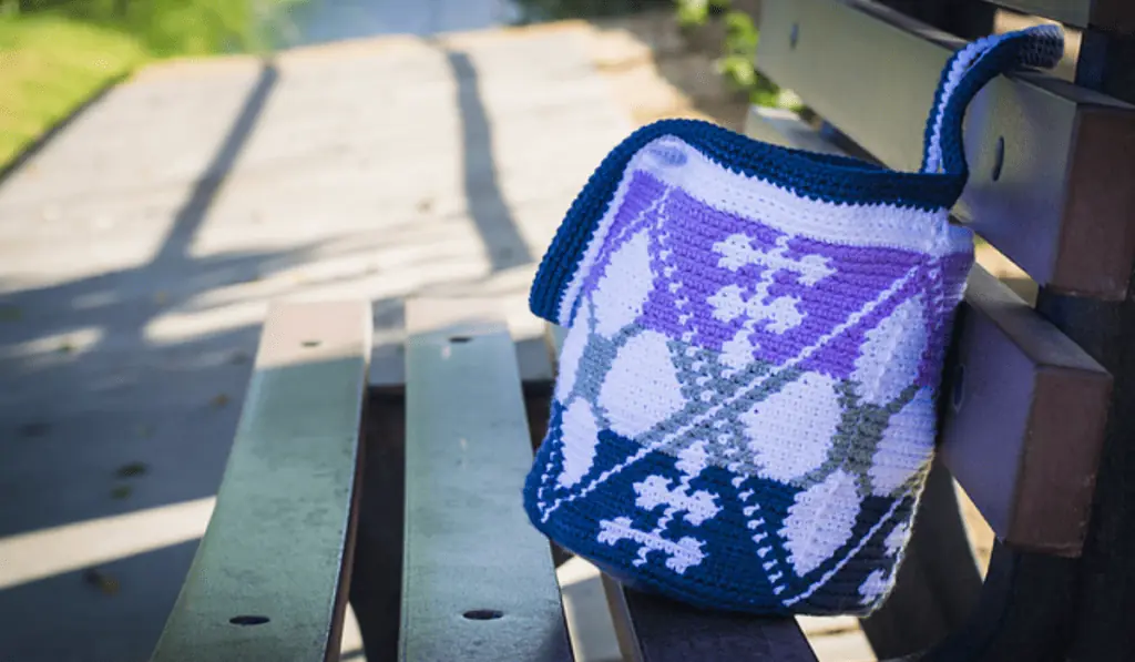 A beach shoulder bag with rows of blue, grey, and purple yarn with white designs on them.