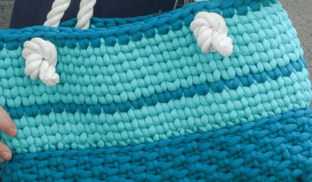 A crochet beach tote with chunky yarn. There are big chunks of dark blue and teal to offset white handles.
