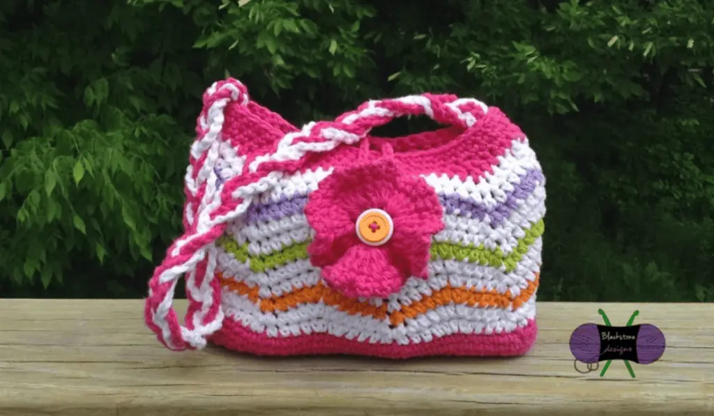 A crochet bag with a hot pink bottom and hot pink top and rows of white, orange, green, and purple yarn.