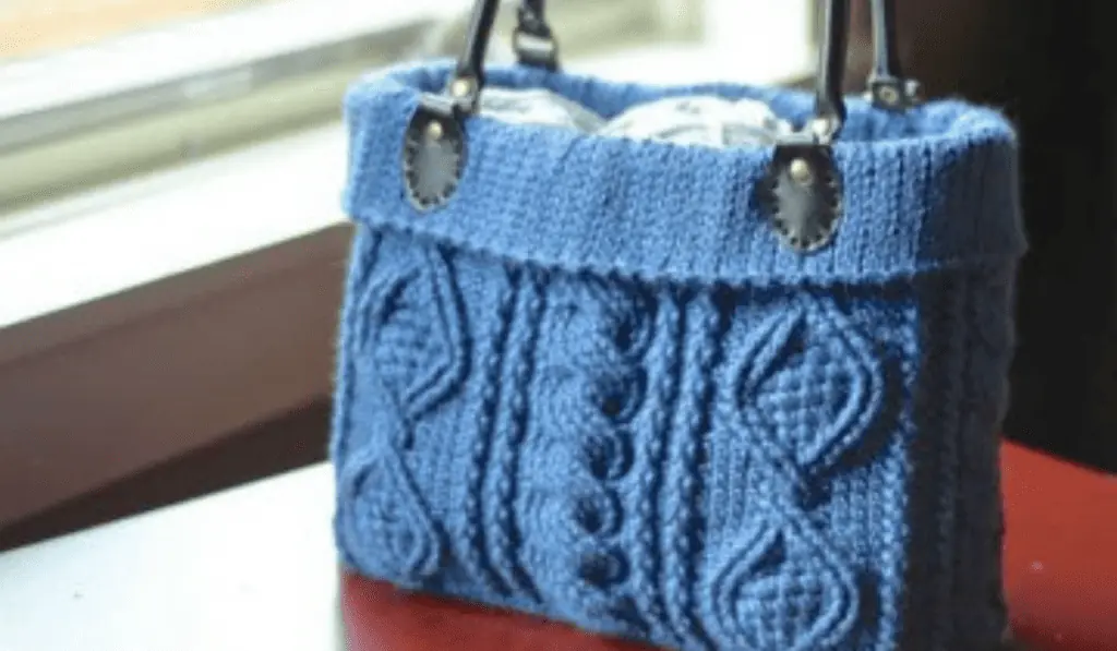 A blue crochet tote bag with leather handles. There are cable designs along the bag.