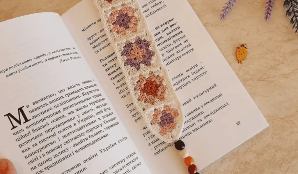 A crochet granny square bookmark with purple, pink, red, and white yarn with wooden beads at the bottom of the bookmark.