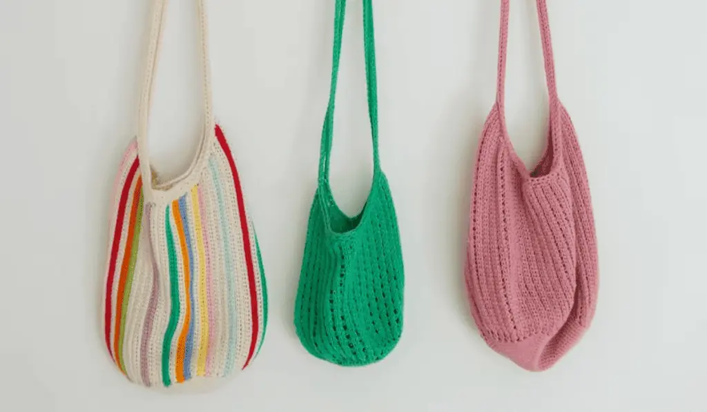 Three crochet tote bags, one is mostly white with rainbow stripes, one is green, and one is pink.