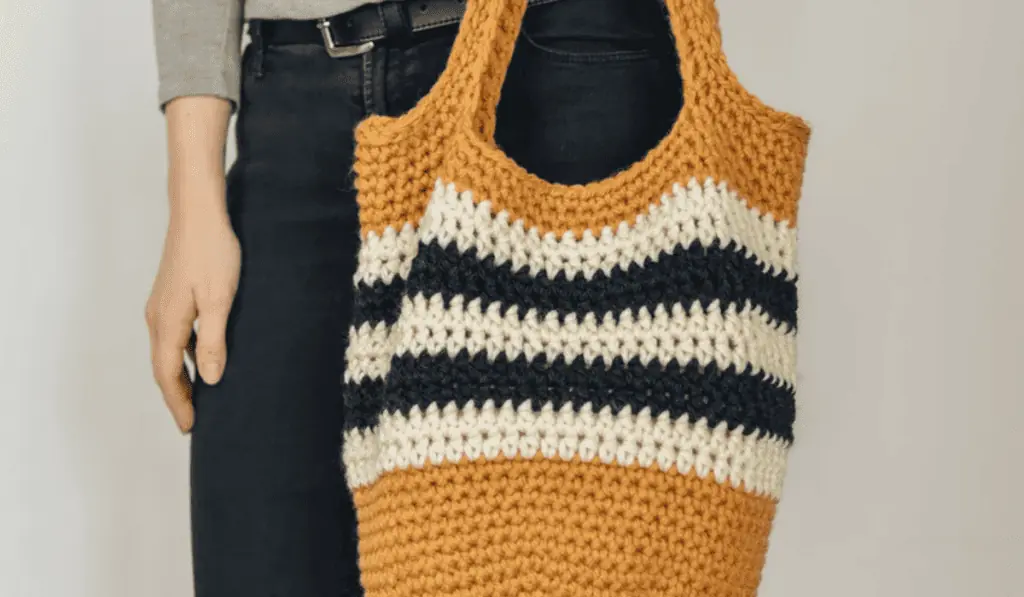 A mostly white crochet tote bag with five lines of color in the middle, three cream and two black.