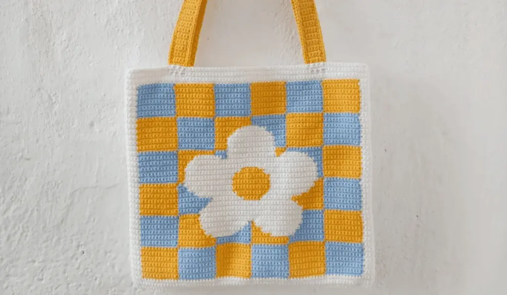 A blue and yellow checkered crochet tote bag with a large flower in the middle.
