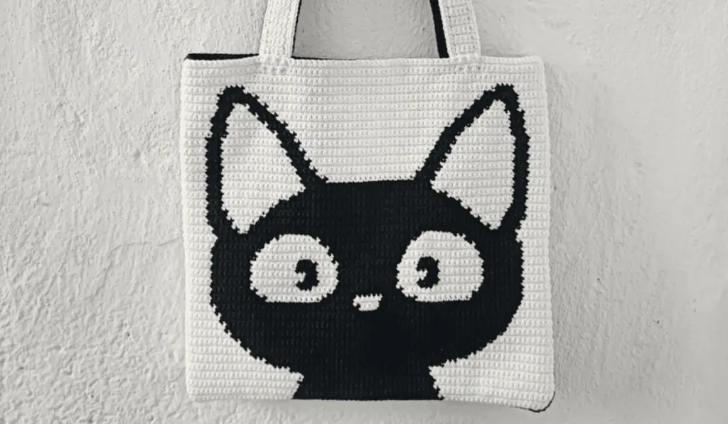 An all white crochet tote bag with a black cat taking up most of the space.