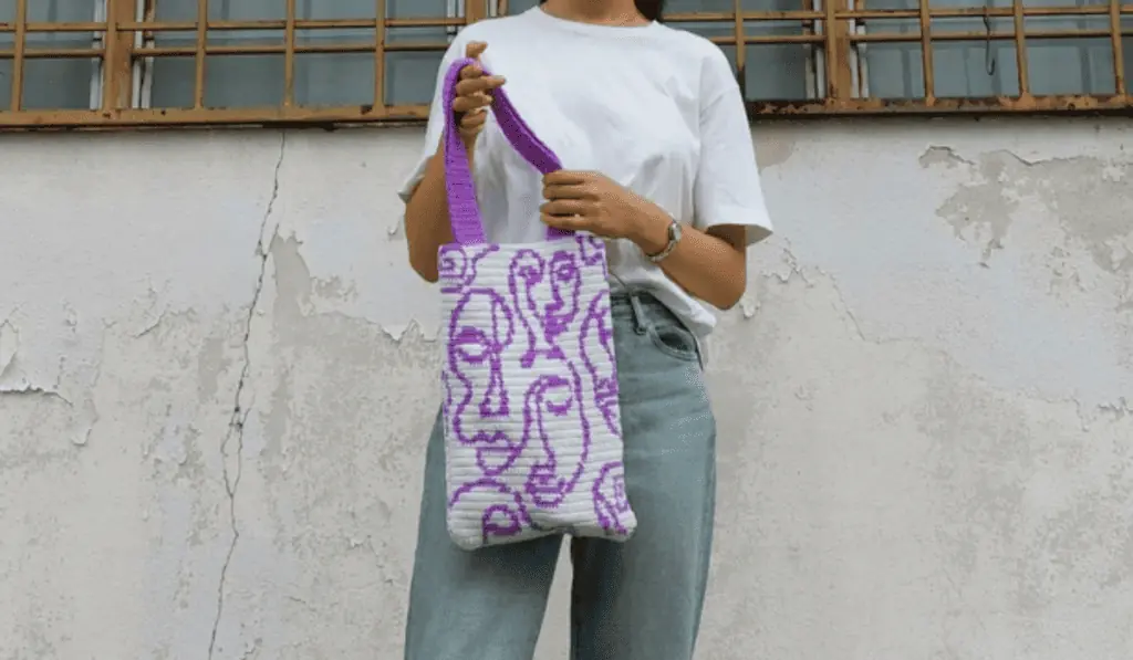 A small crochet tote bag with abstract faces made out of purple yarn on a grey background.