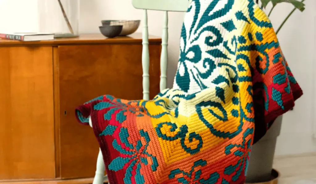 A mosaic crochet blanket patterns that features a background of white, yellow, orange, and red yarn with a dark blue flower design on top.