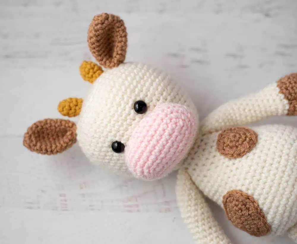 Crochet cow with cream colored body and light brown spots, pink snout and gold horns.