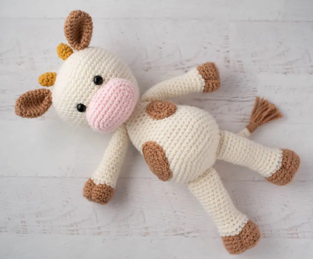 Crochet cow with cream colored body and light brown spots, pink snout and gold horns.