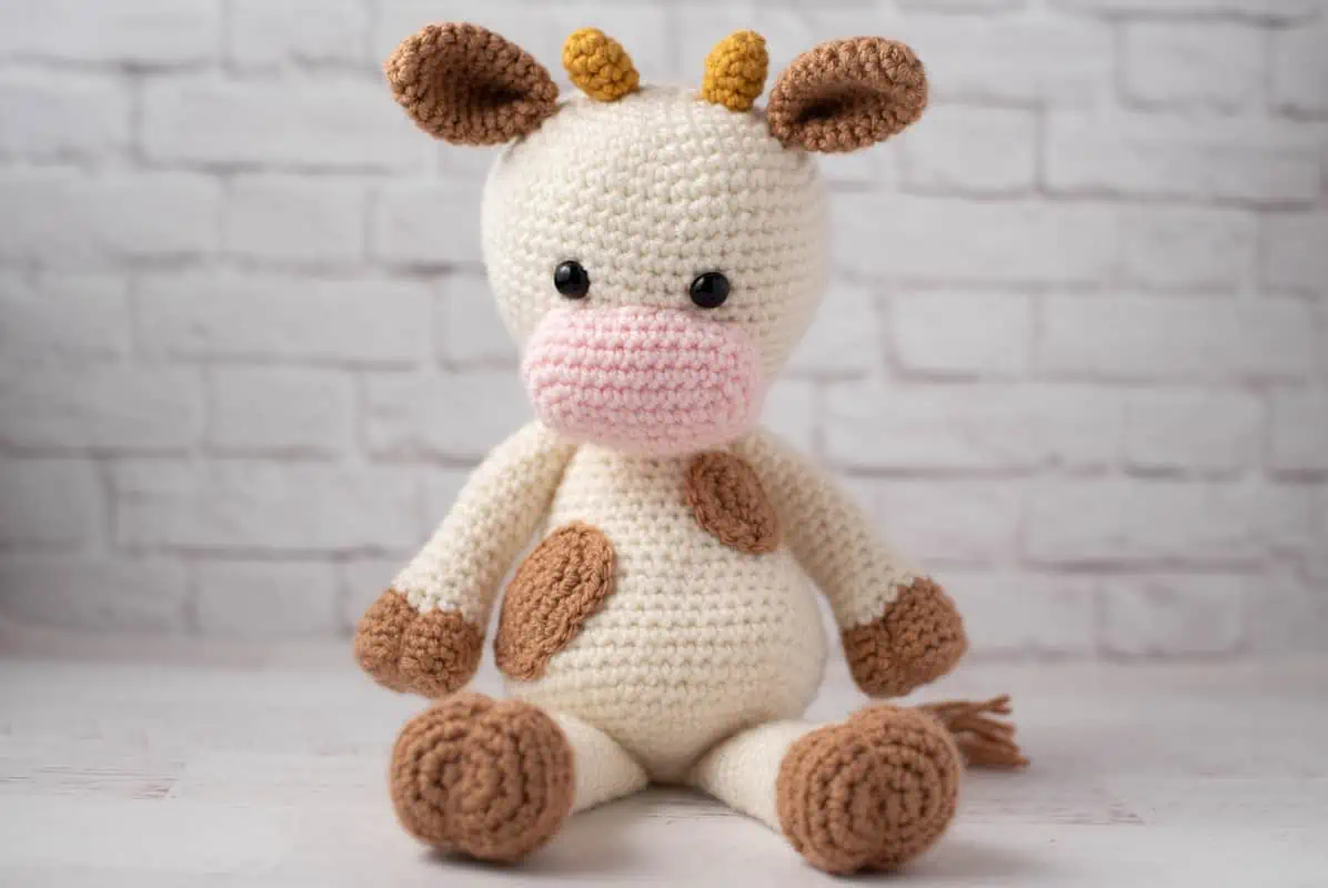Crochet cow with cream colored body and light brown spots, pink snout and gold horns.