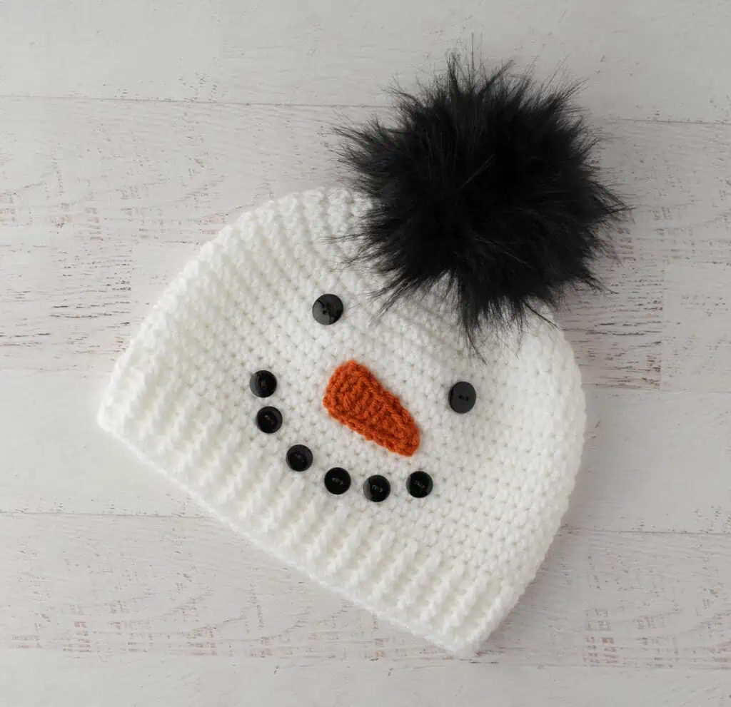 Crochet snowman hat with black buttons, orange carrot nose and black fur pom pom