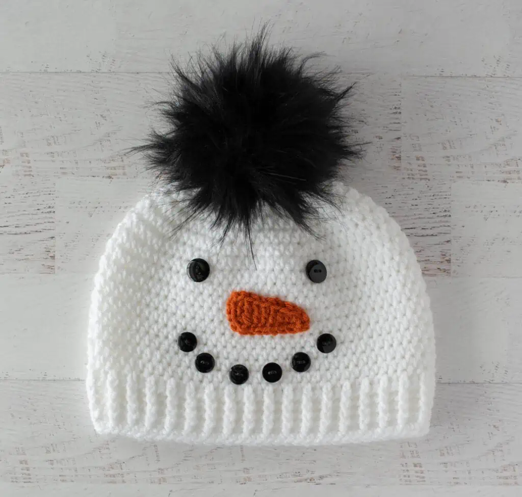 Crochet snowman hat with black buttons, orange carrot nose and black fur pom pom