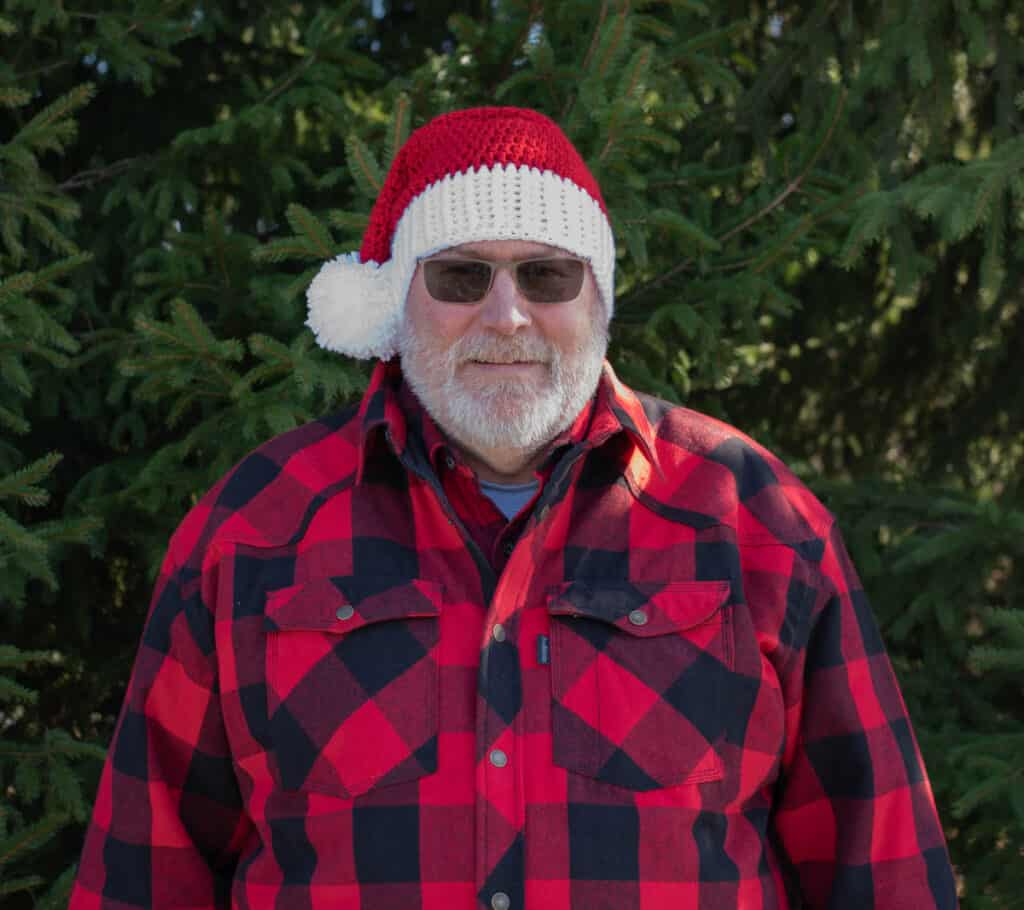 Man in crochet santa hat and red plaid shirt in front of pine tree