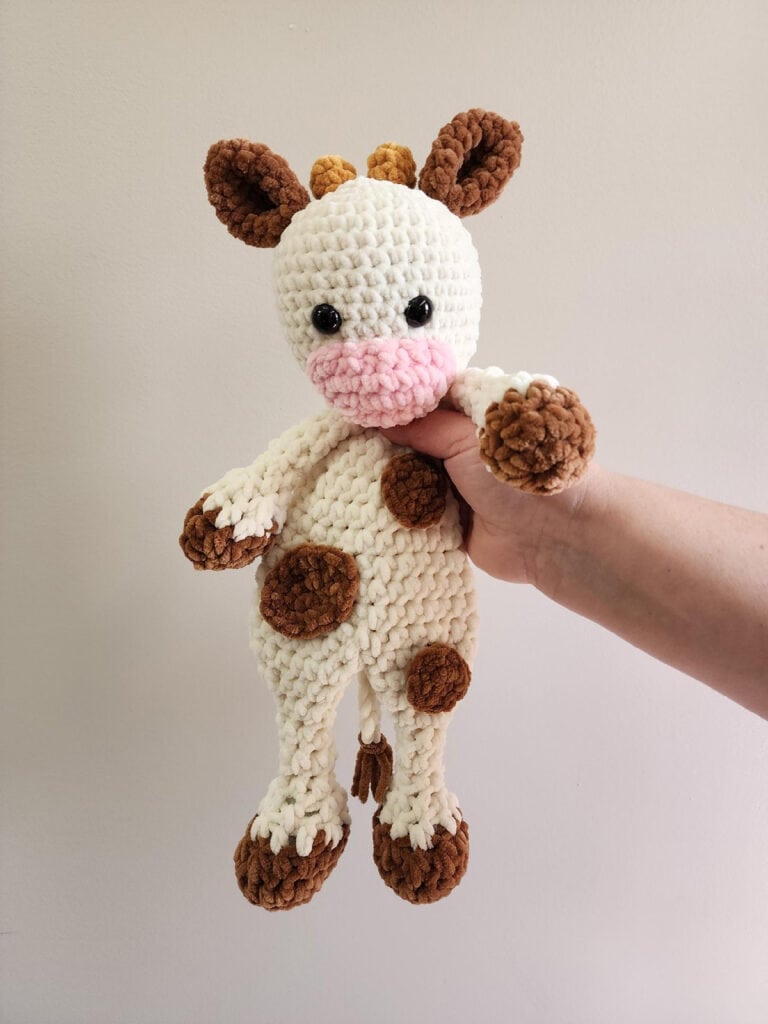 Hand holding crochet cow snuggler made in cream, brown and pink chenille yarn.