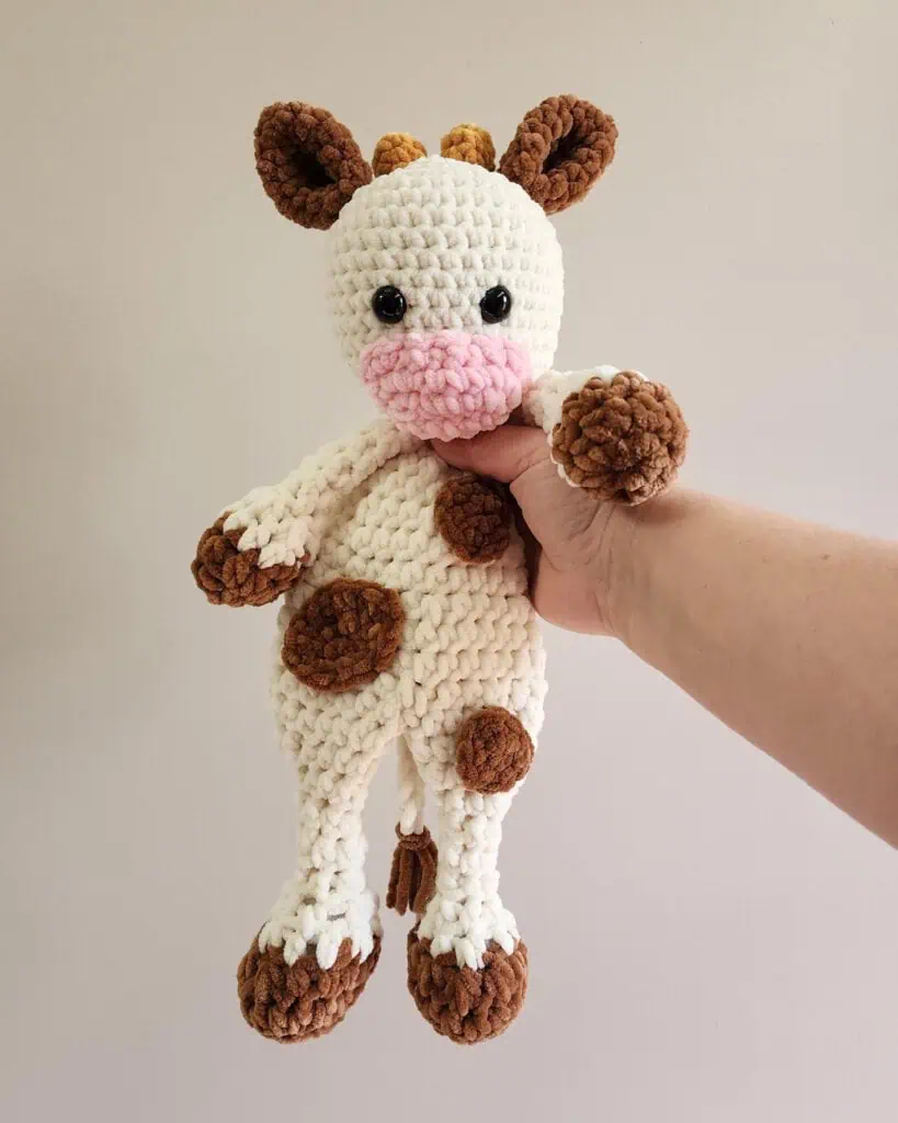 Hand holding crochet cow snuggler made in cream, brown and pink chenille yarn.
