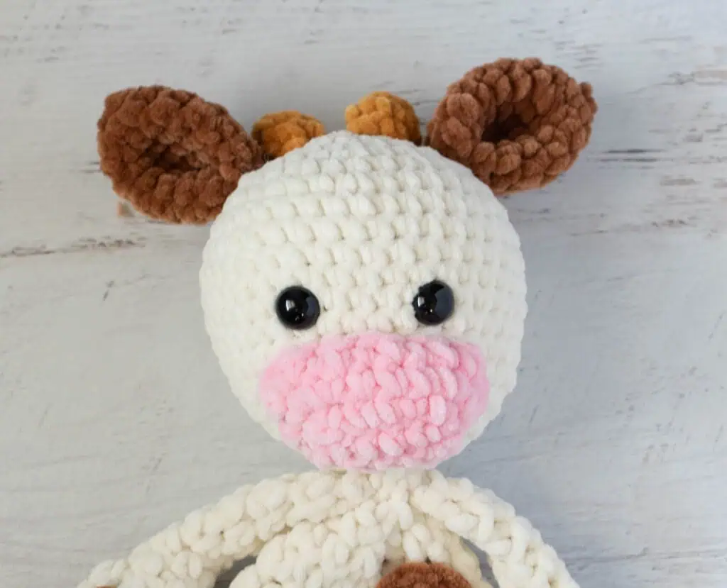 Close up of crochet cow snuggler made in cream, brown and pink chenille yarn.