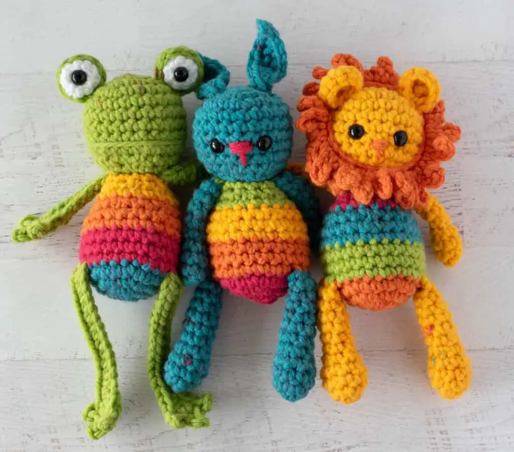 A crochet green frog with rainbow belly, a crochet blue bunny with a rainbow belly and a yellow and orange crochet lion with a rainbow belly