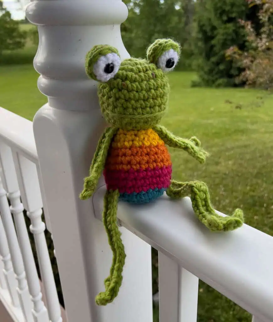 Crochet green frog with yellow, orange, red and blue striped body outside on white porch railing
