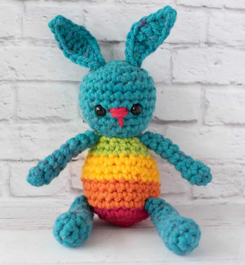 Blue crochet bunny with a rainbow striped body sitting in front of brick wall