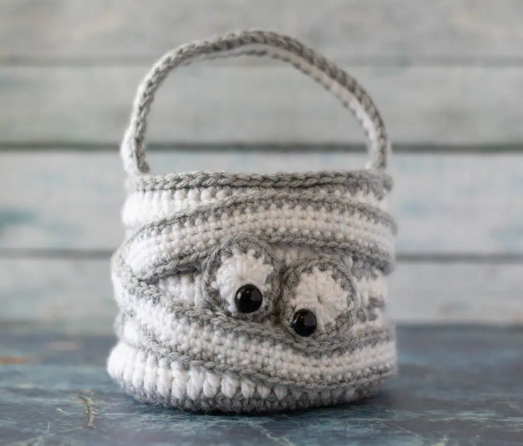 crochet halloween bag in gray and white with straps wrapping bag and eyeballs to look like a crochet mummy.