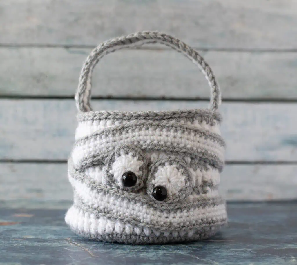 crochet halloween bag in gray and white with straps wrapping bag and eyeballs to look like a crochet mummy.