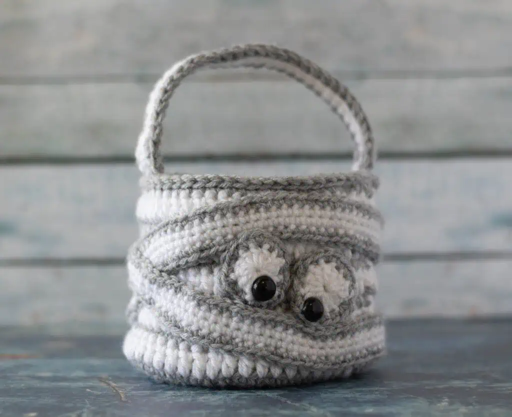 crochet halloween bag in gray and white with straps wrapping bag and eyeballs to look like a crochet mummy.