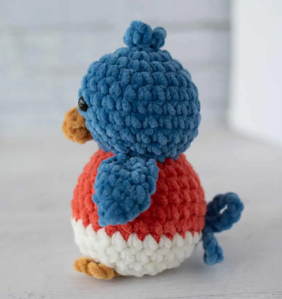 side view of crochet bluebird out of plush blue, mango, cream and gold yarn