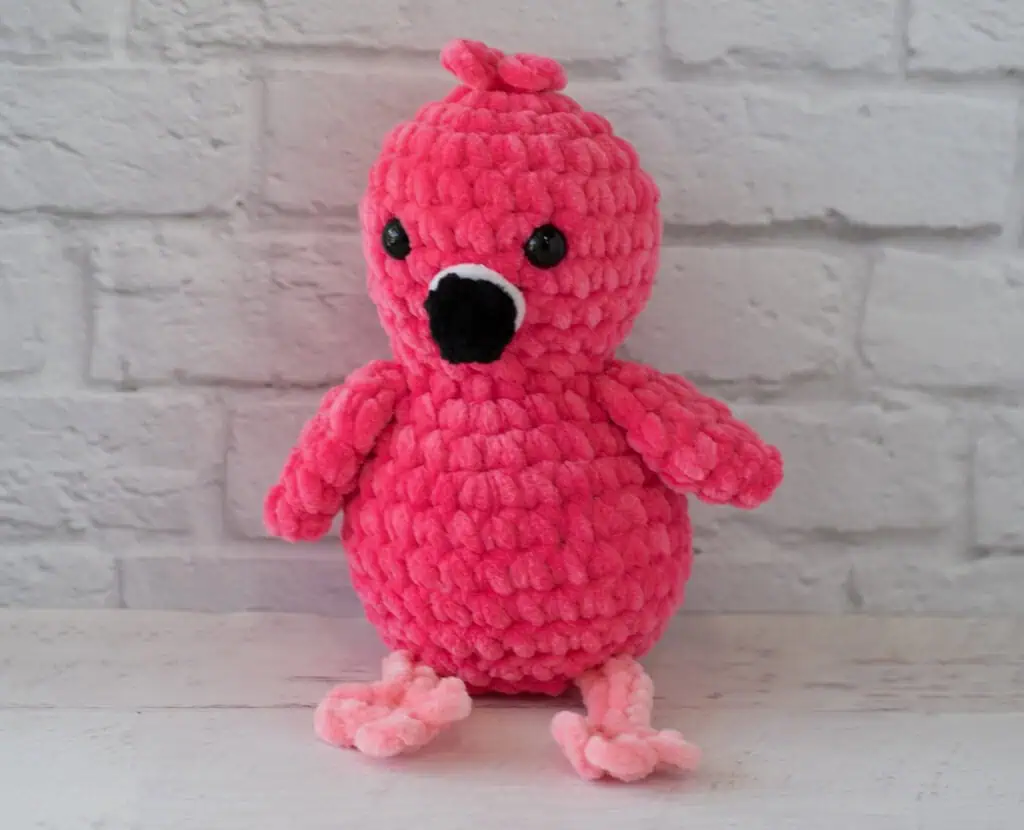 Hot pink crochet flamingo with a black beak and pink legs sitting in front of white brick wall
