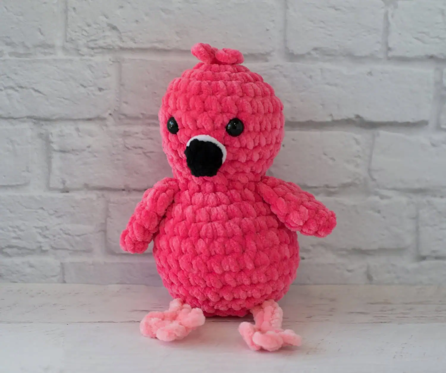Hot pink crochet flamingo with a black beak and pink legs sitting in front of white brick wall