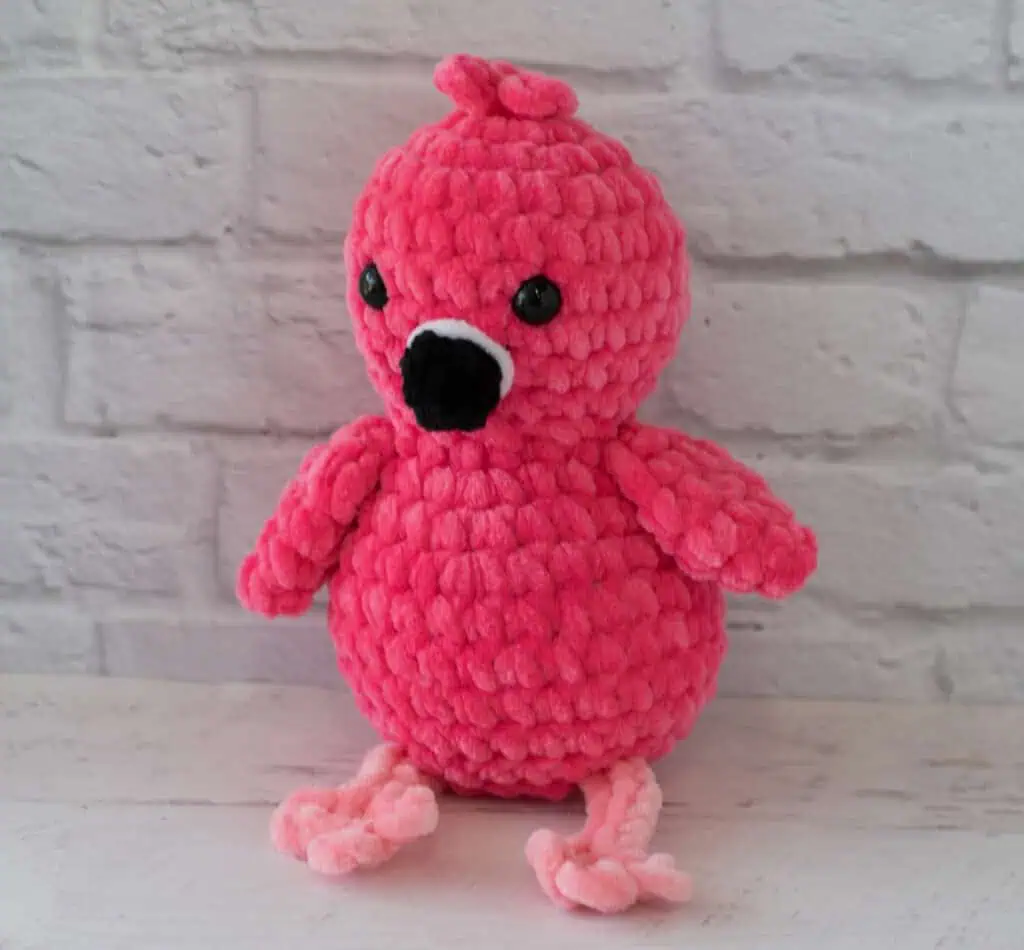 Hot pink crochet flamingo with a black beak and pink legs sitting in front of white brick wall