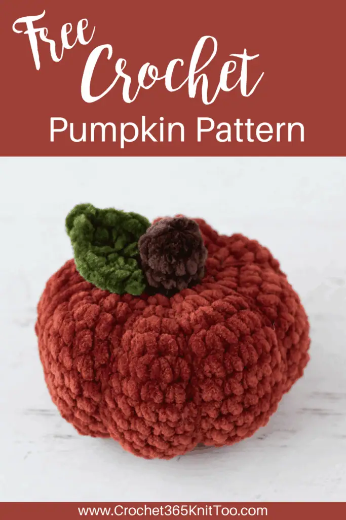 Crochet dark orange pumpkin with green leaf and brown stem out of plush yarn on a white wood table