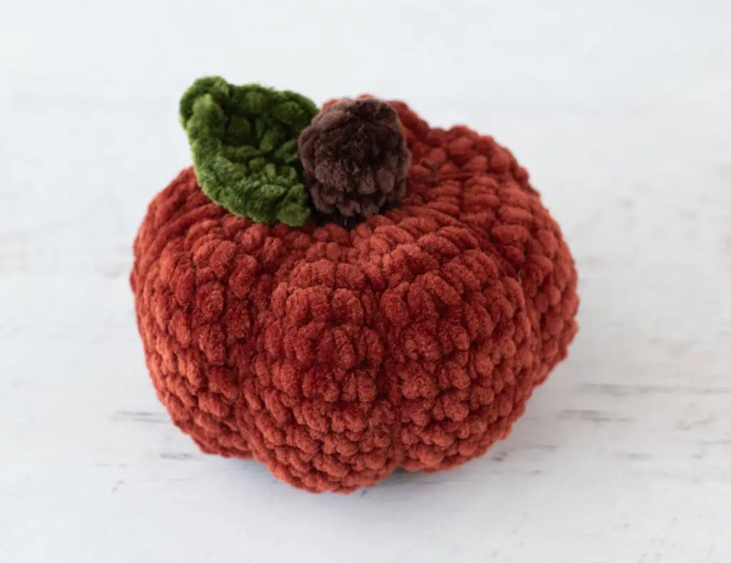 Crochet dark orange pumpkin with green leaf and brown stem out of plush yarn on a white wood table