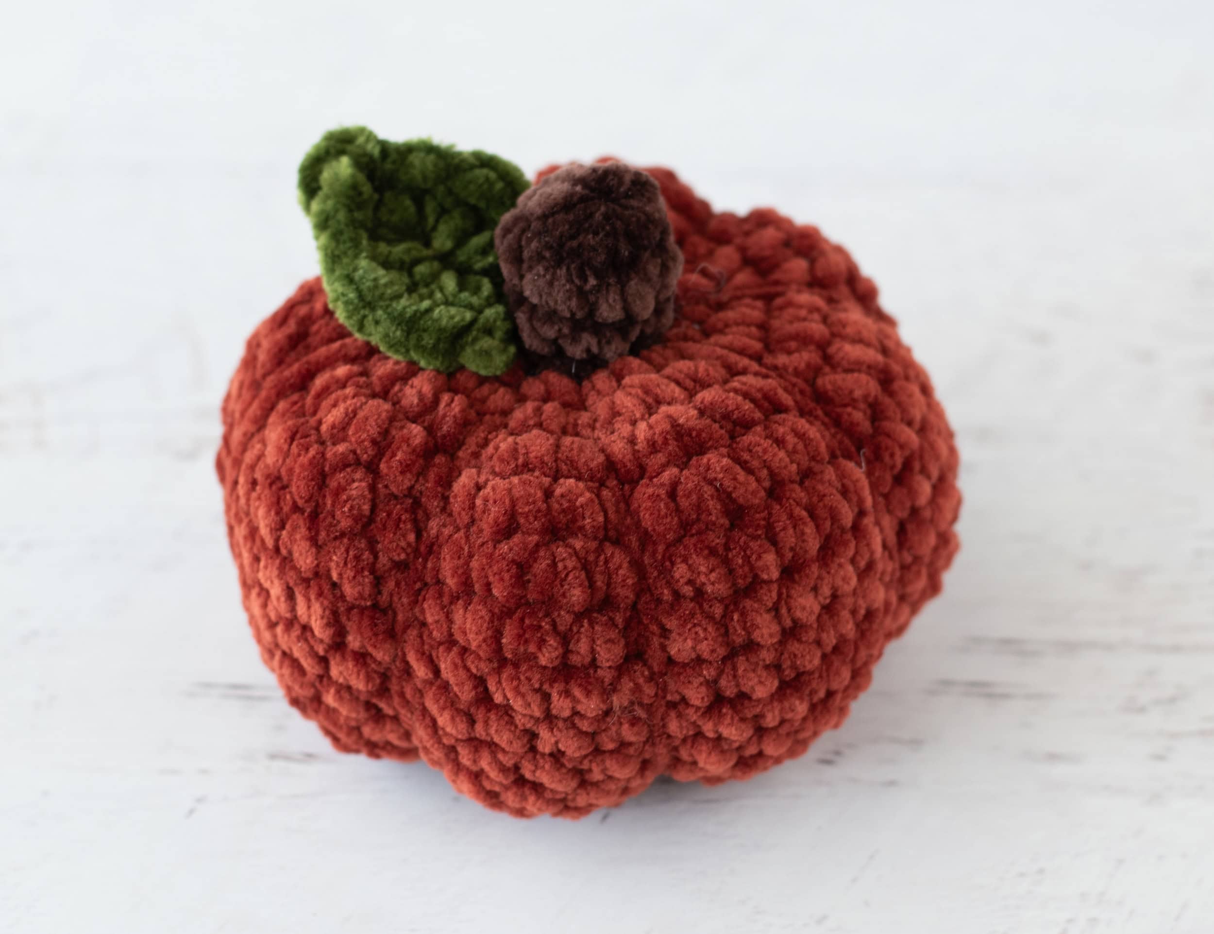 Crochet dark orange pumpkin with green leaf and brown stem out of plush yarn on a white wood table