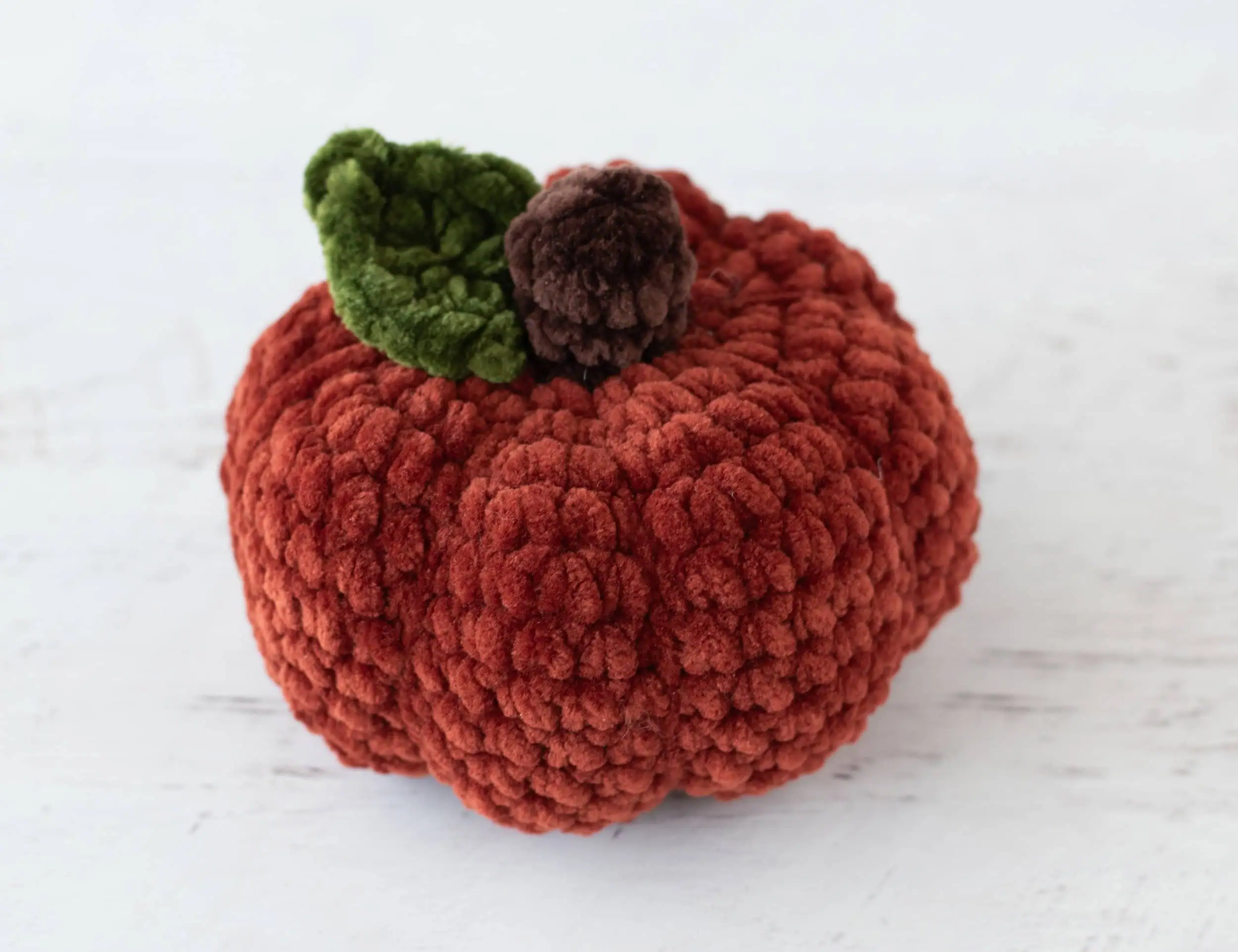Crochet dark orange pumpkin with green leaf and brown stem out of plush yarn on a white wood table