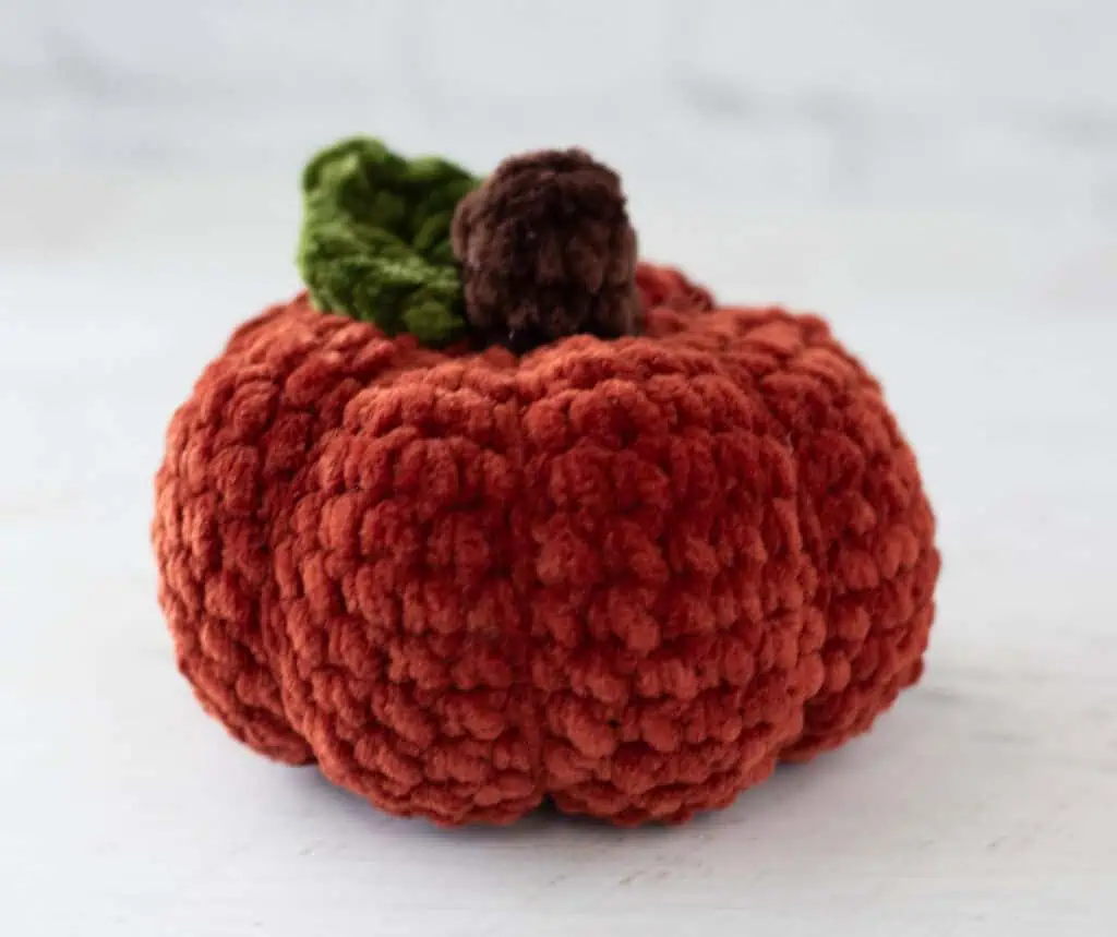 Crochet dark orange pumpkin with green leaf and brown stem out of plush yarn on a white wood table