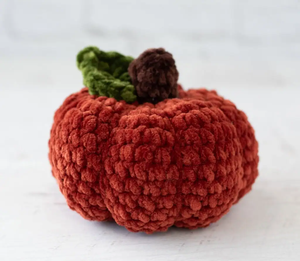 Crochet dark orange pumpkin with green leaf and brown stem out of plush yarn on a white wood table