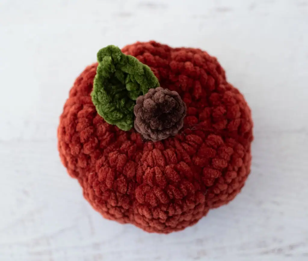 Crochet dark orange pumpkin with green leaf and brown stem out of plush yarn on a white wood table