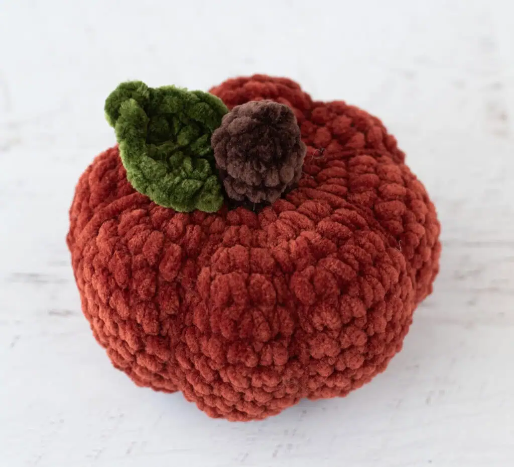Crochet dark orange pumpkin with green leaf and brown stem out of plush yarn on a white wood table