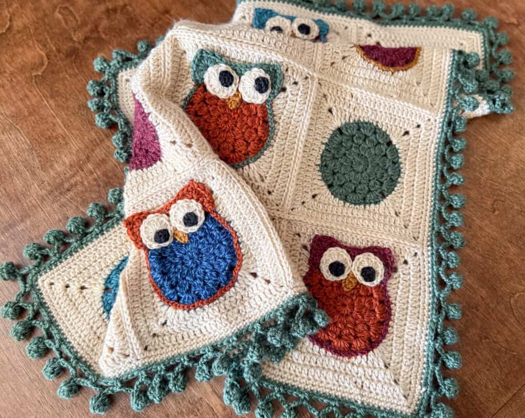 Crochet owl blanket with cream background, red, gold, green and blue owls with a green pom pom border on wood background