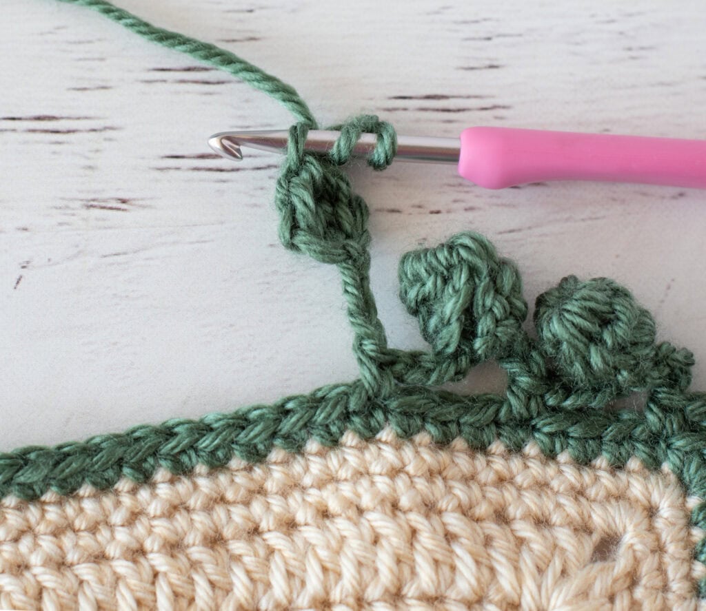 Step by step photo of crochet pom pom border in green yarn on an ivory blanket
