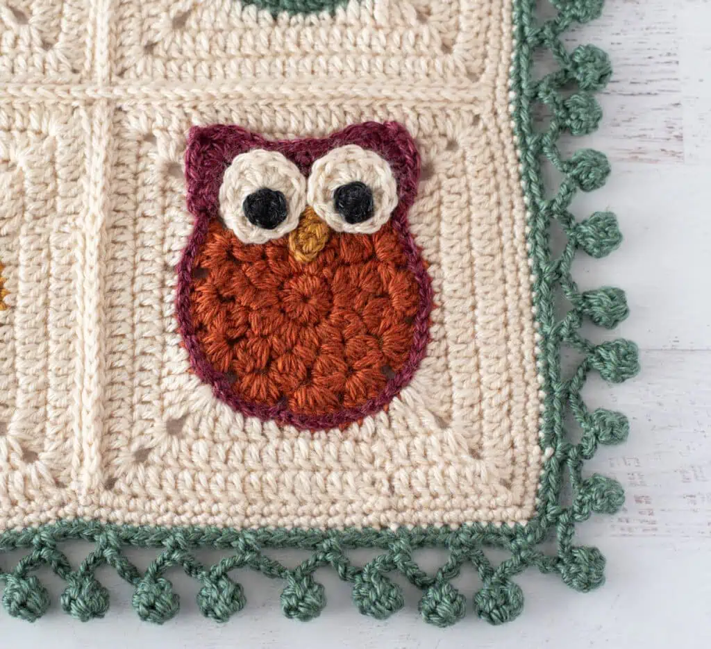 Crochet owl blanket with cream background, red, gold, green and blue owls with a green pom pom border.