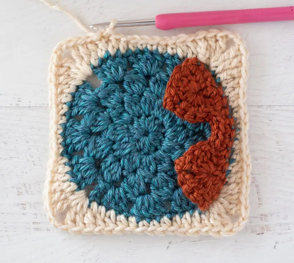 Crochet blue, cream and orange afghan square with pink crochet hook