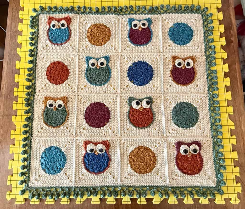 Crochet owl blanket with cream background, red, gold, green and blue owls with a green pom pom border on yellow blocking squares.