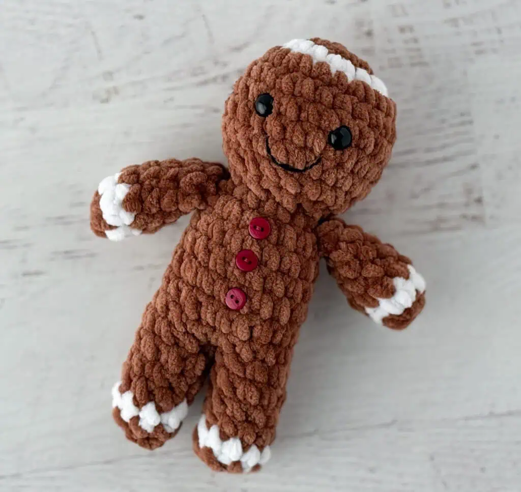 Crocheted plush gingerbread standing on a whitewashed table, features brown and white yarn and red buttons.