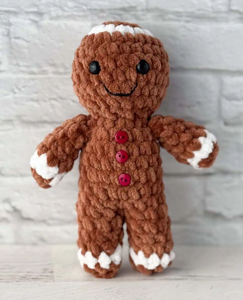 Crocheted plush gingerbread standing on a whitewashed table, features brown and white yarn and red buttons.