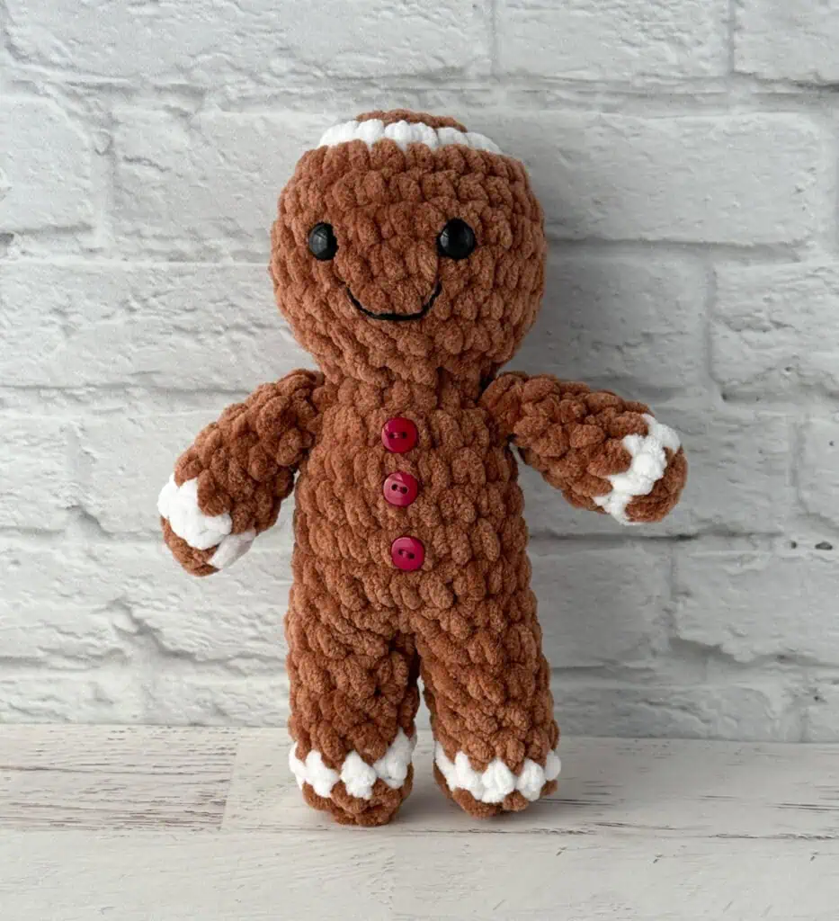Crocheted plush gingerbread standing on a whitewashed table, features brown and white yarn and red buttons.